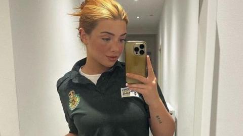 Lorna is using her mobile phone to take a selfie in a mirror here. She has blonde/ginger hair which is pinned up. She's wearing a green Welsh ambulance service polo shirt with a white shirt underneath. 