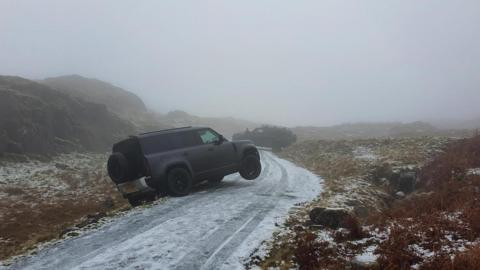 An icy single track road with two black cars run off it. The road is bordered by tall mountains.