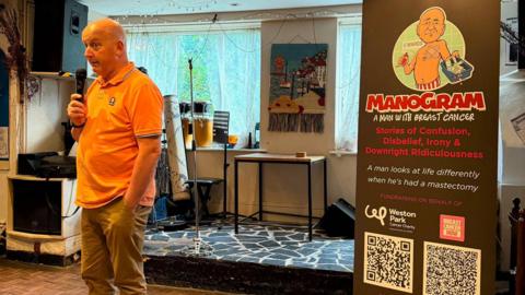 A bald man in an orange polo shirt talks into a hand held microphone