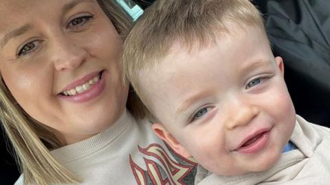 Lydia Collins with fair shoulder-length hair, smiling at the camera while holding her son Rylo, aged about two, with fair short hair and blue eyes' He is also smiling