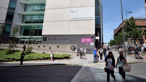 Stock image of people crossing the road in front of the Passport Office, which is a concrete and glass high-rise building