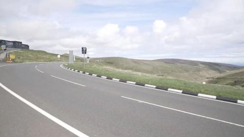 A road in the hills bends round to the right, there is a cafe with black painted walls on the left