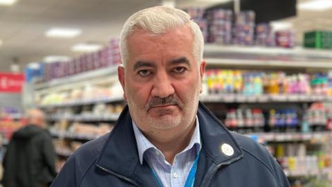 Mark Cox is standing in a store looking directly at the camera. He has short white hair and is wearing a Channel Islands Coop branded coat over a blue checked shirt and a lanyard is visible. To his right are bananas for sale and there are dairy aisles behind.
