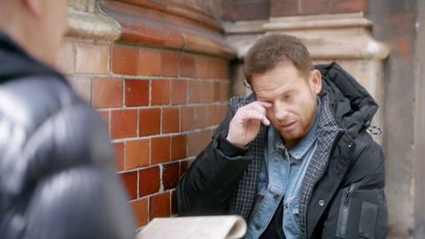 Joe Swash wipes his eye while standing in front of a man holding an old newspaper