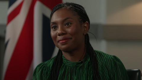 Kemi Badenoch is smiling, and has dark platted hair and is wearing a green and black top. Out of focus behind her is a Union Jack.
