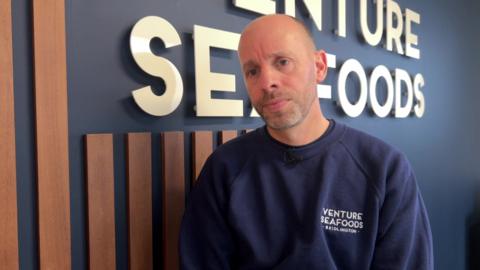 A man with a bald head is standing in his office in front of a wall which shows the business logo of Venture Seafoods. He is wearing a navy blue jumper, which also has the business logo printed on the left side. He's looking pensive as he speaks to camera.