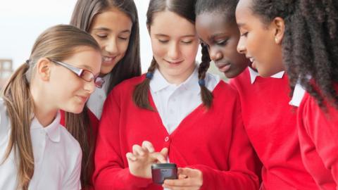 Four young girls crowd around another girl holding smart phone during their school day. They wear red school uniform and are looking excitedly at the screen. 