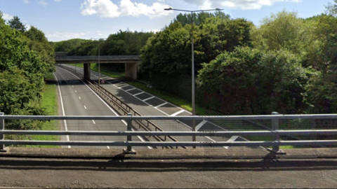 The A442 can be seen from a bridge. There are no vehicles on the carriageways which is has trees on either side.