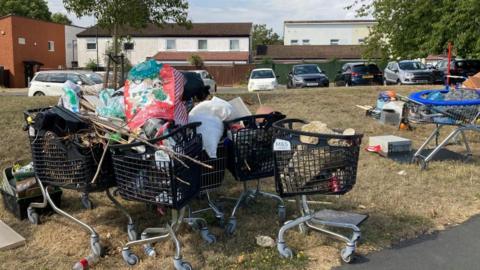 Several black shopping trolleys full waste sit on a patch of grass, there is housing and parked cars in the background. 