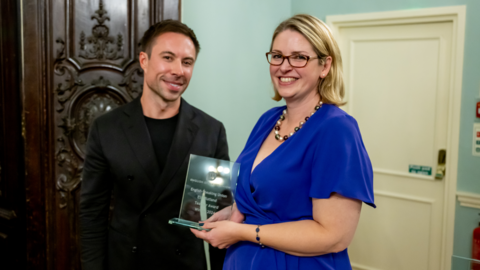 Dr Jo wearing a blue dress holding a glass award next to  the presenter in black shirt and black jacket 