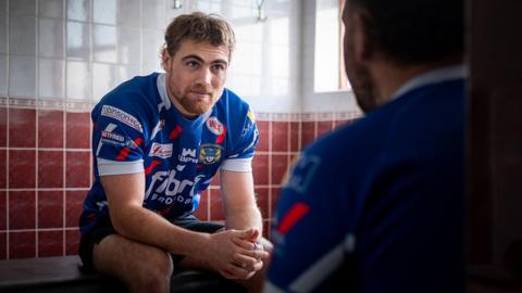 Zarrin Galea, wearing his Workington Town kit, sits on a bench in the club's changing room as he talks to another player who is sitting opposite. Galea has brown hair and a beard.