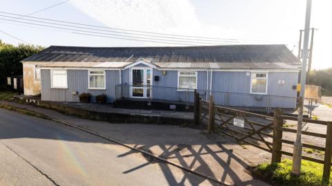 Exterior of the single-storey property. The village hall has five windows and a glass entrance. A wooden gate leads into the property. It has an accessible slope which leads to the entrance of the building.