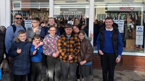 The photo shows a group of people smiling at the camera. They are all standing in front of a shop, which reads 'St. Anne's Charity Shop' in bold black letters.