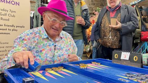 Gerald Whitehouse wearing glasses, a bright pink hat and a colourful shirt. He is sat at a blue table playing backgammon. The table has round pieces on it with red and yellow arrows. There are two people stood watching the player on the right. One is wearing a black and gold waistcoat and is holding a takeaway coffee in his right hand.