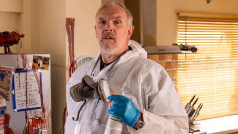 The image shows an indoor kitchen setting featuring a person standing in the foreground, wearing protective cleaning equipment. The person is dressed in a white disposable coverall suit, along with bright blue protective gloves. Around the neck is a grey respirator mask with dual filters, hanging loosely against the chest. In one hand, the person holds a spray bottle of cleaning solution.