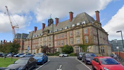 An external view of Peacock Hall which is part of the Royal Victoria Infirmary (RVI). The building is made of red brick in a similar style to grand French houses and has ivy growing up the walls. Cars are parked outside. Six of the building's chimneys can be seen. In the middle of the roof stands the lantern toped with a copper dome and weather vane.