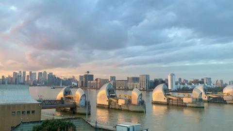 The sun glints off the Thames flood barriers and the sky is full of blue, grey clouds