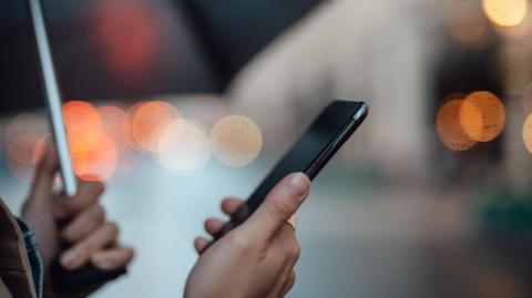 A person holding an umbrella looking at a mobile phone with lights, which are out of focus, in the background 