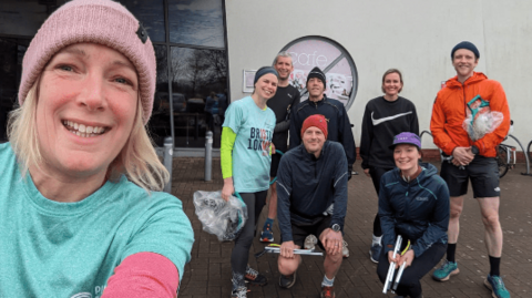 A selfie with a close-up of Claire, smiling to camera, and seven other ploggers dressed for running and holding plastic bags and litter sticks