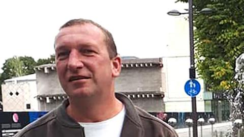 A head and shoulders shot of Robert Kvederis who has short brown hair and is wearing a brown jacket over a white T-shirt. Behind him a building can be seen - possibly a civic centre.
