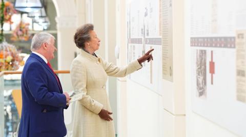 The Princess Royal viewing the Liberation Trail exhibition during a visit to St Peter Port. She is pointing at one of the boards and a man standing slightly behind her appears to be explaining it and he is holding a programme in his hand. She is wearing a long cream coat with brown gloves and he is wearing a navy suit with a red tie.