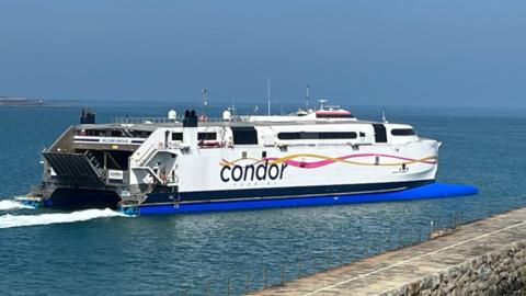 Condor ferry moving through the water near a harbour. The ferry is white. Its Condor logo is printed down the side of the ferry.