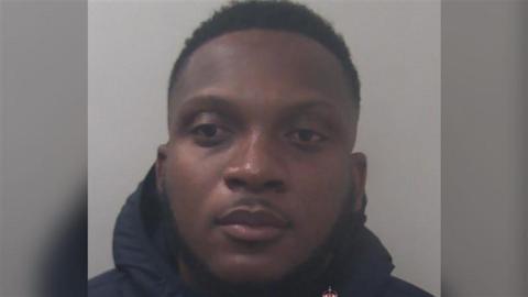 A mugshot of a man in a navy hoodie. He is in front of a grey background.