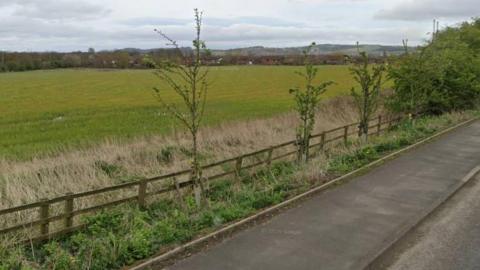 A general view of the green field where the homes would be built. It is next to a road, with a housing estate visible in the distance.
