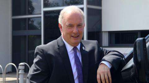 A man with short white hair leans on a van while wearing a dark-coloured suit with a purple tie.