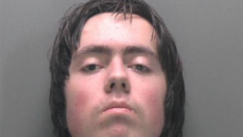 A young man is staring into the camera. He has dark brown long hair, dark brown eyes and is stood in front of a dark grey background.