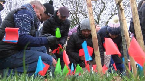 People plant tiny blue, red and green flags in the ground. About five people are seen planting the flags while wearing warm clothing.