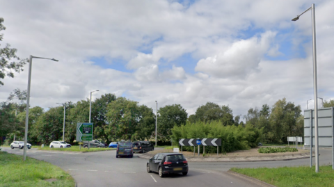 A roundabout with green bushes in the centre and a number of cars driving around