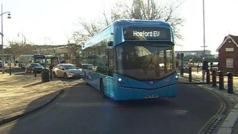 A blue bus turning a corner in the road pulling out of a bus stop, with Hoeford EV written on the front of it.