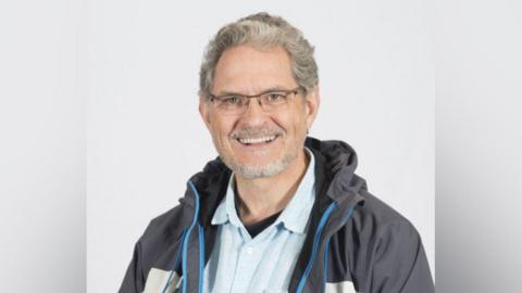 Councillor Brian Wernham. He has short grey curly hair and trimmed beard. He wears glasses, a light blue shirt and and outdoor grey and blue jacket. The head-and-shoulders portrait was taken against a white background. 