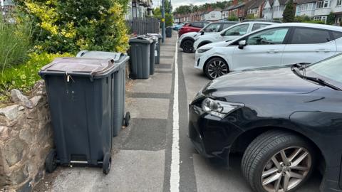 A wide residential street, with a rows of cars parked on its right, all coming up to a white line. On the left, a line of wheelie bins by a wall further restrict the space for people to pass.