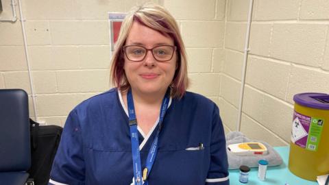 Fay is in the middle of the picture, wearing blue nurses scrubs and a blue lanyard. She's looking at the camera and wearing glasses. She's sat in a white, breezeblock room.