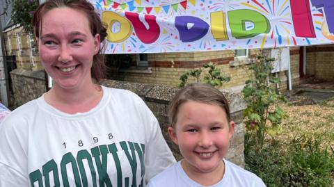 Ms Sired and her daughter are standing in front of a white banner which reads "you did it". She has brown hair that has been pulled back and is wearing a white top with 'Brooklyn 1898' printed in green on the front. He face is red after completing the challenge with her daughter. Evie is smaller than Ms Sired and is also wearing a white top and has slightly flushed cheeks. She is also smiling.