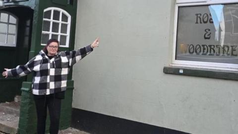 A woman standing in front of the entrance to a pub. She is wearing a black and white coat and black trousers. She has her arms out to the sides as she is celebrating becoming the pub's landlady.