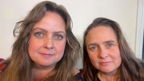 two green-eyed women looking into the camera. They are identical twins and although they have styled their long hair differently, the similarity in their faces is still evident