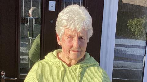 A woman with white hair and wearing a yellow top standing outside a front door. It is a sunny day and there are shadows in the background.