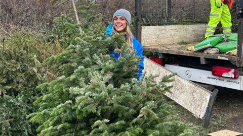 A woman with long, blonde hair wearing a grey beanie and blue coat standing behind a Christmas tree.