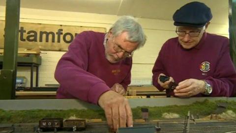 Two men in burgundy jumpers are leaning over a model railway display intently as they check the track and hold black items to go on it. A workshop can be seen on the wall behind them.