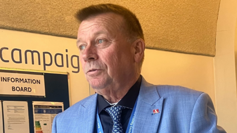 A man in a blue suit and tie looking to the side in a hallway with a notice board on the wall behind him.