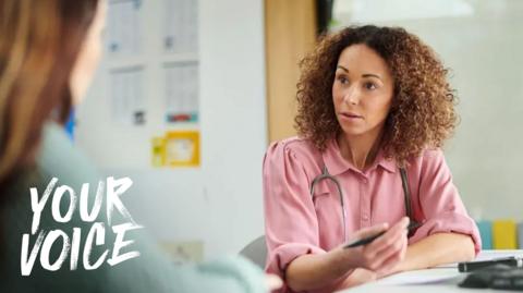 A posed picture of a GP appointment - a woman wearing a pink shirt and with a stethoscope round her neck is sat at a desk talking to another woman who is out of focus on the left of the shot. The woman in the pink shirt has brown curly hair and is holding a pen in her hand.