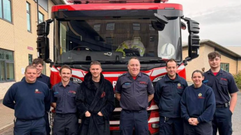 Eight people are standing in front of a red fire engine. A building is on the left of the photo and a smaller building is in the background on the right.