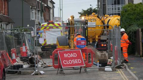 Workmen in hi-vis have fenced off a road. there is a Road Closed sign in the foreground and yellow tankers are behind the fenced off area.