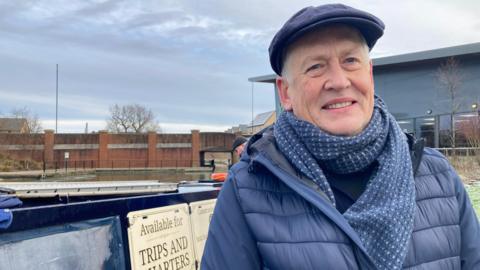 Mike Gibbin is standing on the towpath. There is a barge behind him. He is wearing a blue jacket and a scarf.