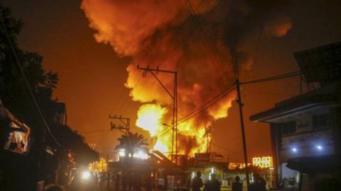 A cloud of fire and smoke rises over a cluster of buildings in the background at night, turning the sky dark orange