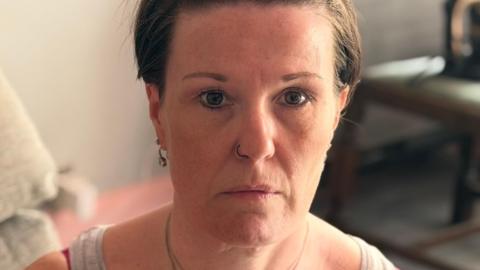 Lee-Ann Hawkes, who has short brown hair, sits on a sofa looking ahead with a serious expression. 
