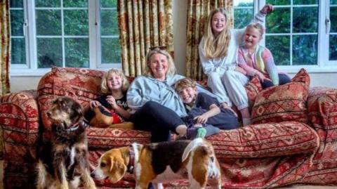 Mylo and his family in a living room, on a sofa in front of windows and patterned curtains: Mylo cuddles his mum, who are sat on the sofa next to one of Mylo's siblings. Two of his other siblings sit on the back of the red patterned sofa. Two dogs are in front of the sofa, one is sat down and looking to the left, while the other, a beagle, stands and looks to the left